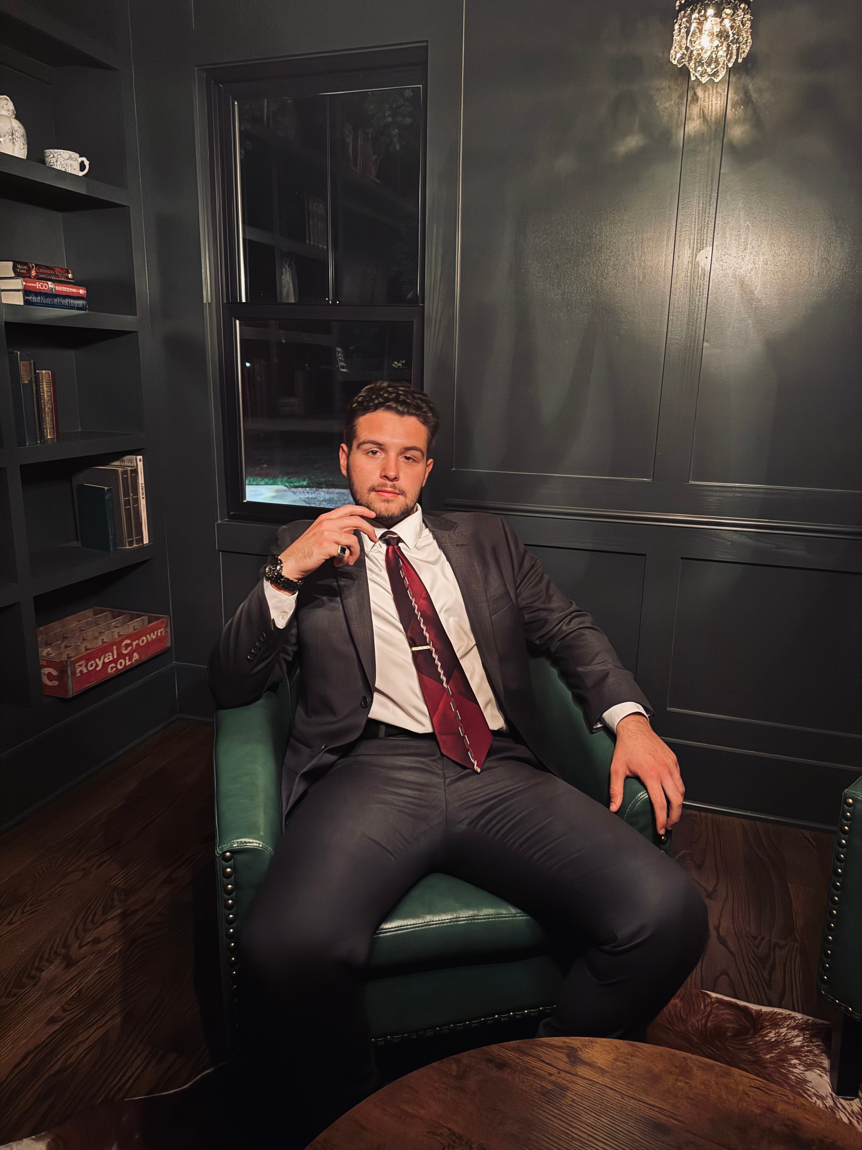 Alex Gruich, a young entrepreneur with short dark hair, wearing a smart-casual shirt, stands confidently in a modern Akron office with large windows and tech devices visible. The background features subtle blue and teal tones, reflecting a professional yet approachable atmosphere.
