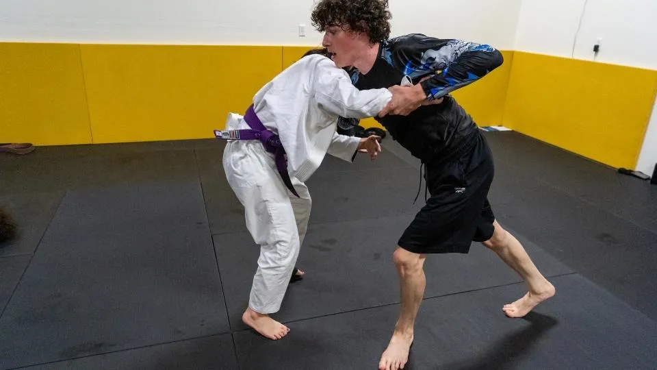 Adult Brazilian Jiu-Jitsu class at Mountain Rollers Jiu-Jitsu in Colorado Springs featuring both gi and no-gi training. A purple belt student in a white gi engages in stand-up grappling with a no-gi student, demonstrating versatility and skill development in different BJJ styles. Mountain Rollers Jiu-Jitsu offers comprehensive training for adults, focusing on both gi and no-gi techniques to enhance self-defense skills and fitness in Colorado Springs.