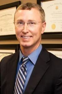 Dr. Craig Meier in a suit coat, standing with arms relaxed and smiling in a bright clinic hallway with diplomas and natural lighting.