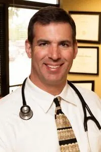 Dr. David Allen smiling in a white coat, seated in a clinic setting with framed medical credentials in background.