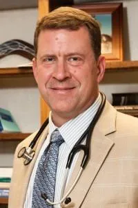 Dr. David Sharp in a lab coat, seated confidently with a relaxed smile in a modern clinic, framed by books and framed credentials.