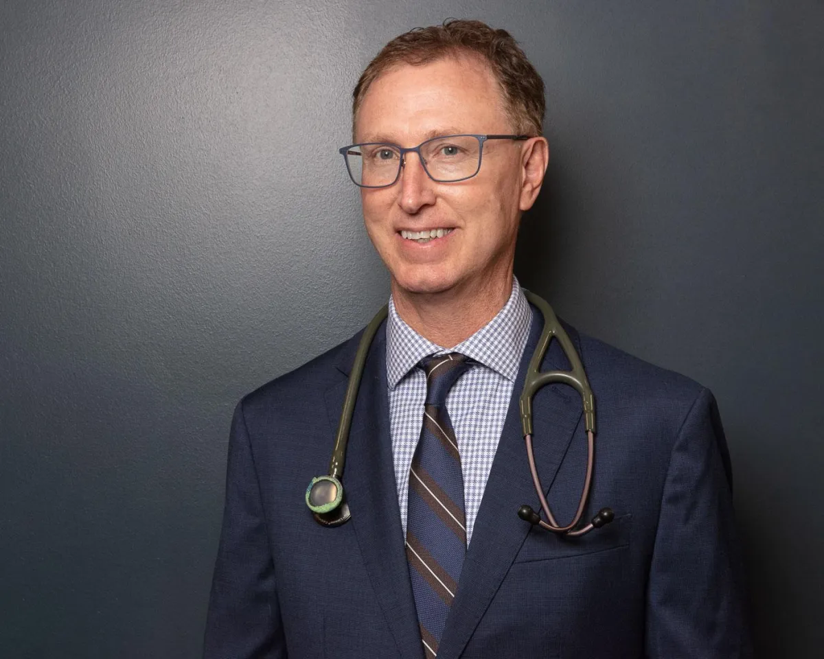 Dr. Craig Meier in a suit coat, standing with arms relaxed and smiling in a bright clinic hallway with diplomas and natural lighting.
