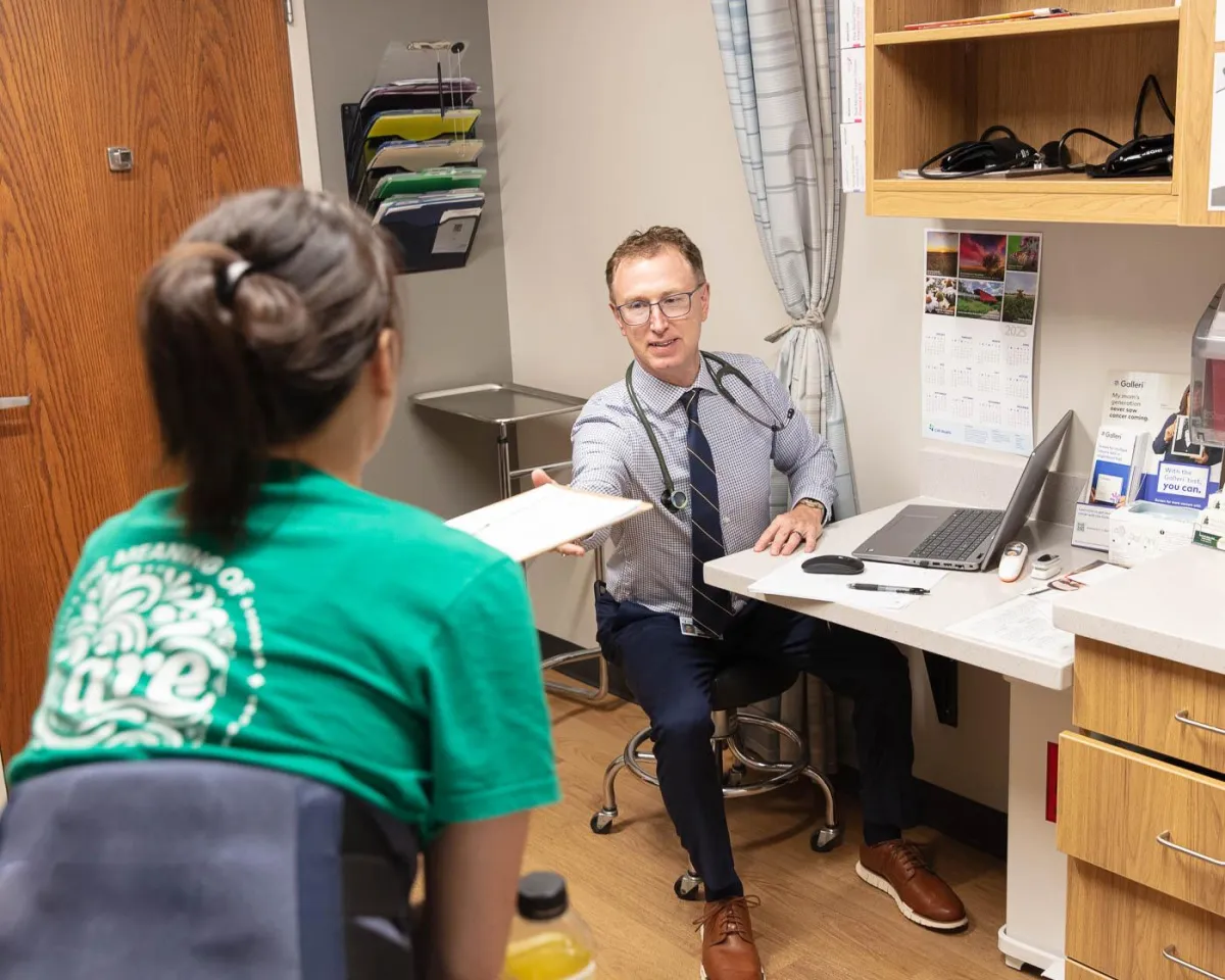 A professional photograph of an examination room in Omaha, showcasing the Chronic Care Management Services offered. The image captures a male doctor, sitting at his desk, and a patient, who is in focus, receiving a clipboard from him. The doctor, who appears to work for the patient, is wearing a stethoscope and a tie, reflecting a caring and dedicated approach. The room is tidy, with medical supplies, and features a calendar and a laptop on the desk, illustrating a modern and efficient practice.