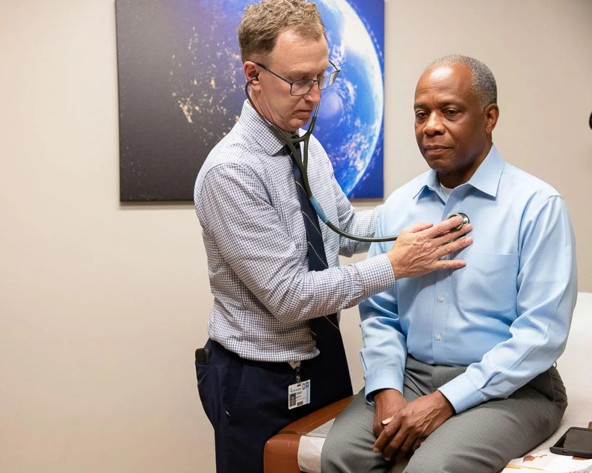 A male doctor, dressed in a collared shirt and tie, is attentively listening to the chest of an older male patient with a stethoscope. The patient is seated calmly on an examination table, looking forward. Behind them, a large framed image of Earth from space hangs on the wall, adding a professional yet calming backdrop to the clinic room. This image effectively communicates the focused and personalized approach to annual physicals available in Omaha, highlighting the importance of regular monitoring and dedicated medical attention for ongoing health conditions.