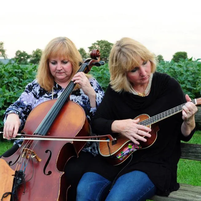Laurie Reese, cellist, and Tish Brown, ukulele, sit on a backyard bench playing music