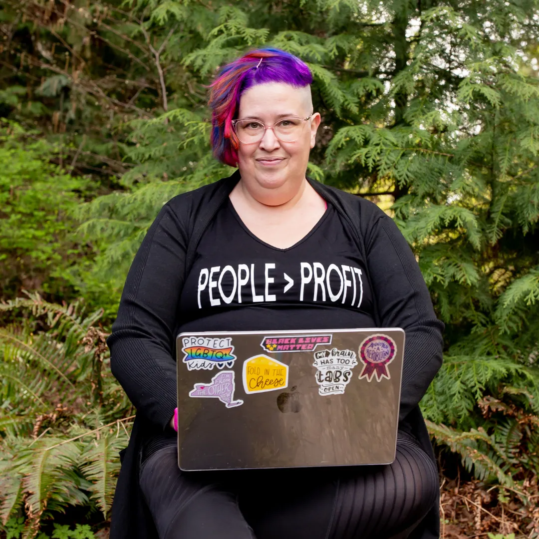 Meg Brunson smiles while working outside on her laptop. Her hair is purple and pink and her shirt says "people > profit"