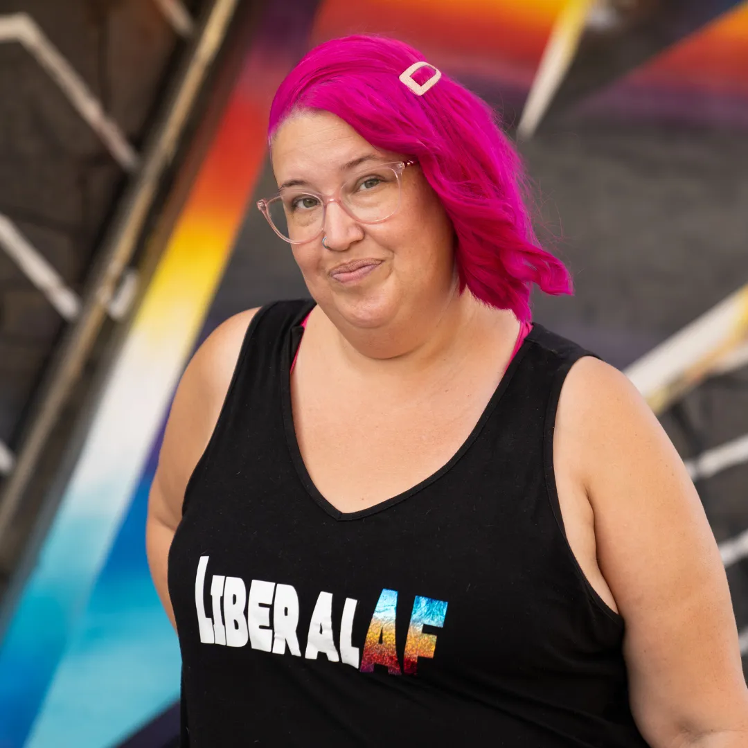 Meg is smiling at the camera. She wears a "LiberalAF" tank top and has hot pink hair.