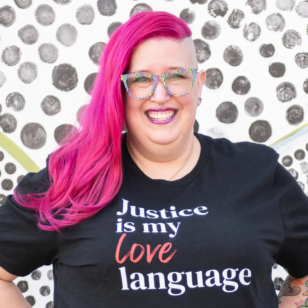 Meg Brunson is smiling with her hot pink hair pulled to one side. She wears a black shirt that says 'Justice is my love language,' and rainbow leopard print glasses.