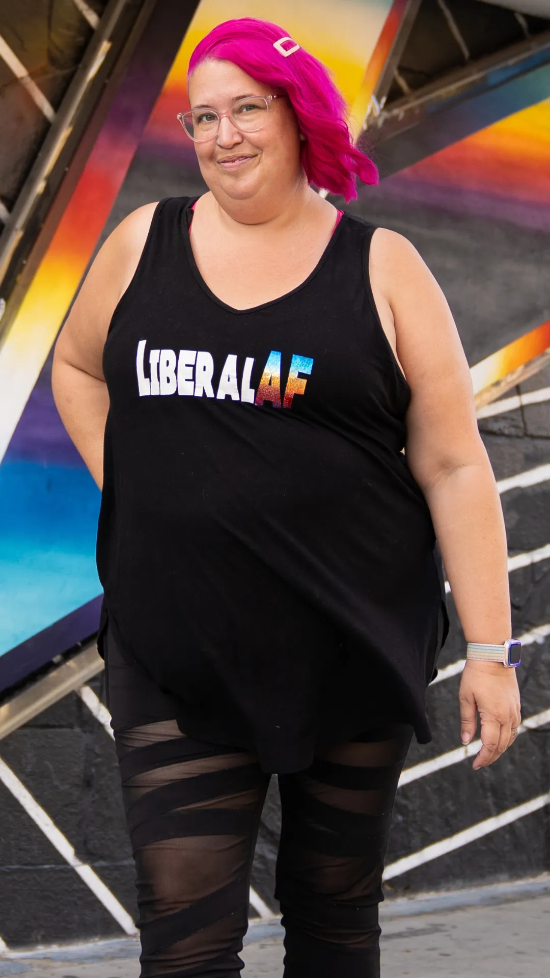 Meg is standing and smiling at the camera. She wears a "LiberalAF" tank top and has hot pink hair.