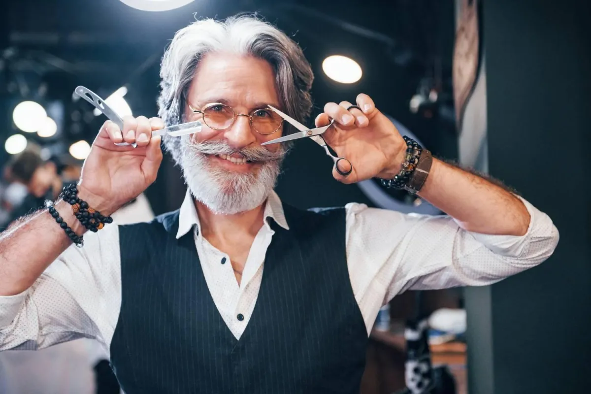 Homme souriant avec une moustache stylisée tenant des ciseaux et un rasoir.