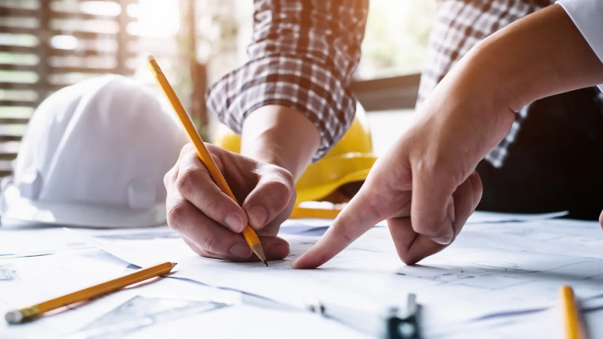 Close-up of two people collaborating on construction blueprints with pencils, planning a building project. A white hard hat and yellow helmet are visible in the background, emphasizing the design and planning phase of construction work.