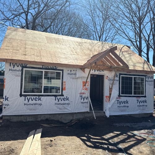 New ADU under construction with Tyvek house wrap, exposed roof framing above the front entry, and installed windows. The detached structure is in the framing and exterior sheathing stage of the building process.