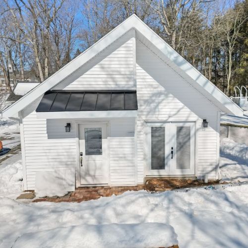White detached ADU with a steep gable roof, black metal awning, and modern exterior lighting, surrounded by snow-covered landscaping and leafless trees in a winter setting. Large glass patio doors provide natural light and a contemporary design aesthetic.