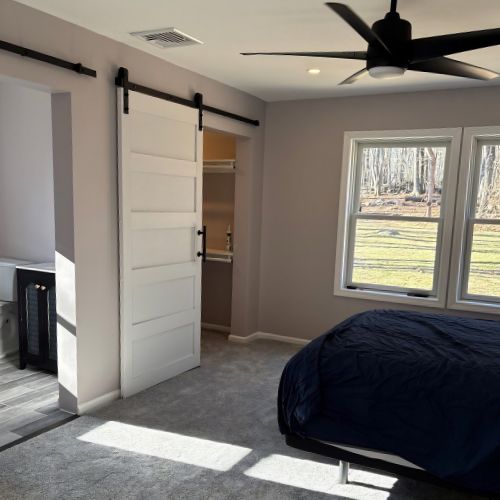 Modern ADU bedroom with a sliding barn door closet, large windows providing natural light, neutral-colored walls, and a ceiling fan. The room features soft carpeting and a bright, open layout with views of a wooded backyard.