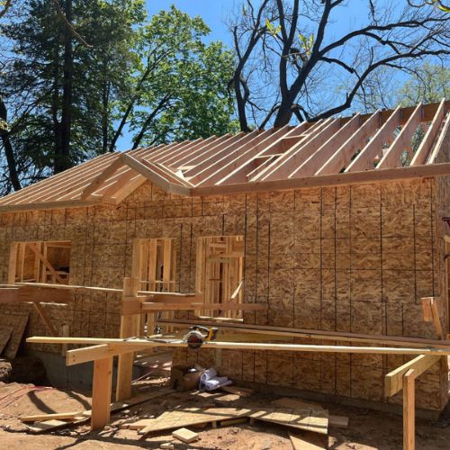 ADU under construction with wood framing, OSB wall sheathing, and exposed roof rafters at a residential job site surrounded by trees.
