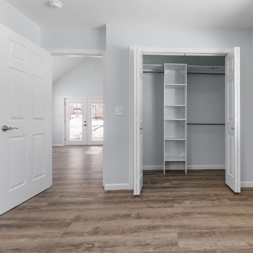Bright, modern ADU interior featuring wood-look flooring, light gray walls, and an open closet with built-in shelving. Double doors lead to a spacious living area with natural light streaming through large glass doors, showcasing a clean and functional layout.