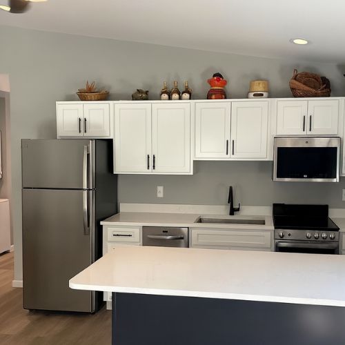 Modern ADU kitchen featuring white shaker cabinets, stainless steel appliances, a built-in microwave, and a large island with a white countertop. The bright, open-concept space includes gray walls, wood-look flooring, and contemporary finishes.