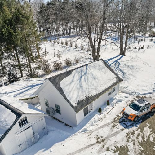Aerial view of a detached white ADU surrounded by snow-covered landscaping, trees, driveway access, and a nearby work truck in a residential backyard.