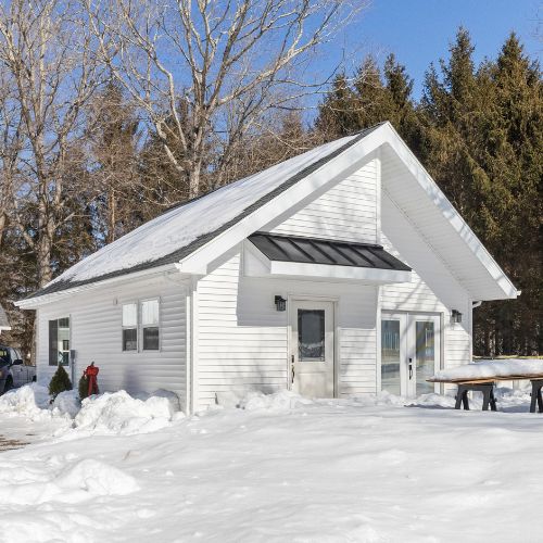 White modern accessory dwelling unit (ADU) with a gable roof and black metal awning, surrounded by snow-covered ground and tall trees on a clear winter day. Large sliding glass doors and clean exterior finishes create a bright, contemporary appearance.