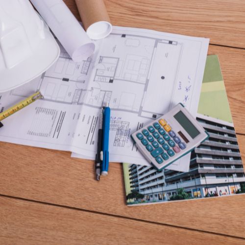 Architectural plans with a calculator, pen, measuring tape, hard hat, and building photo on a wooden table.
