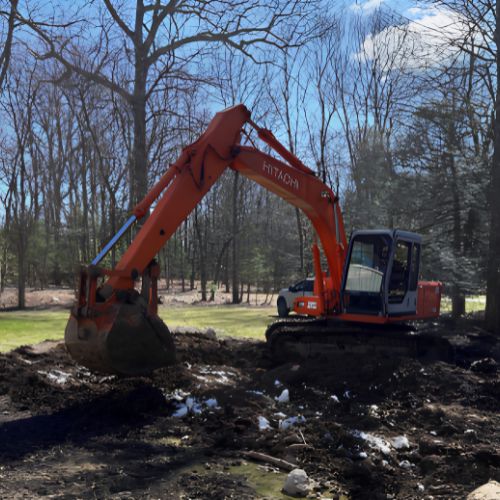 Excavator clearing and grading land for an ADU construction project, with disturbed soil and construction activity visible in a wooded residential area.