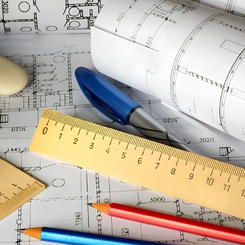 Blueprints with colored pencils, a yellow ruler, a blue eraser, and rolled architectural plans spread across a work surface.