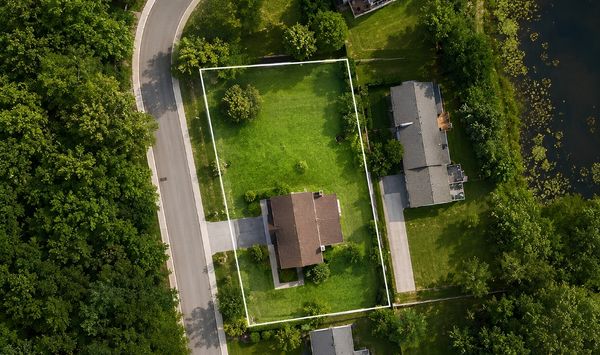 Aerial view of a residential lot outlined in white, showing a home, lawn, driveway, nearby road, surrounding trees, and a pond.