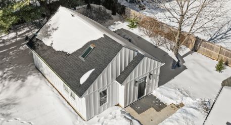 Aerial winter view of a detached ADU with a snow-covered roof, skylight, black entry door, and cleared walkway.