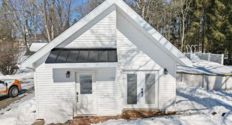White detached ADU in winter with a steep gabled roof, black metal awning, two exterior doors, and snow-covered surroundings.