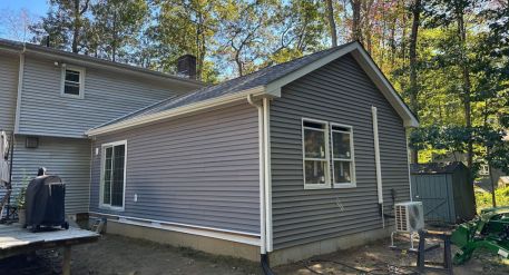 attached ADU addition with white trim, large windows, and an exterior HVAC unit beside a wooded backyard. 
