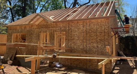 Wood-framed ADU under construction with exterior sheathing, roof rafters, window openings, and a worker on a lift.