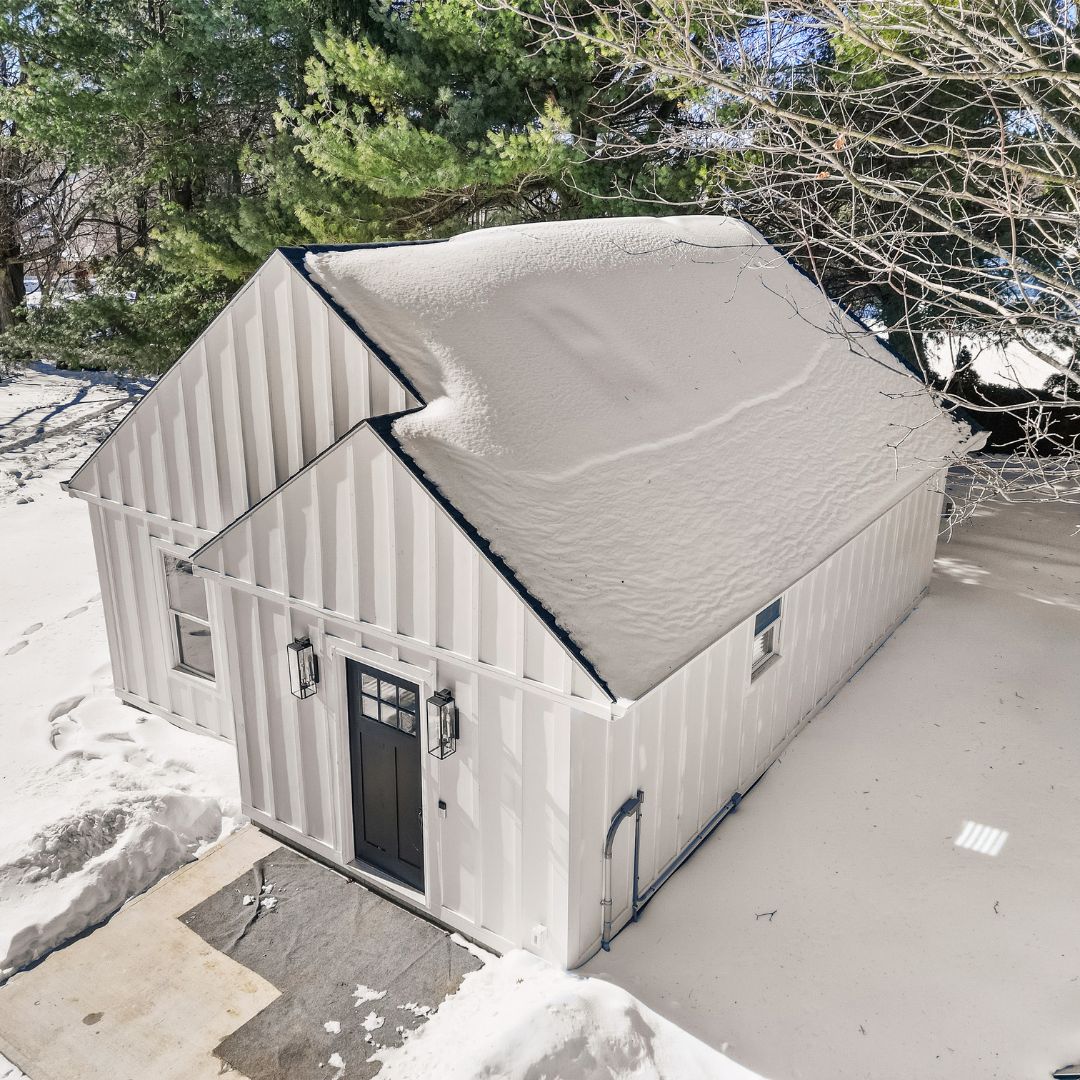 Aerial winter view of a white detached ADU with a snow-covered roof, black entry door, and cleared front walkway.