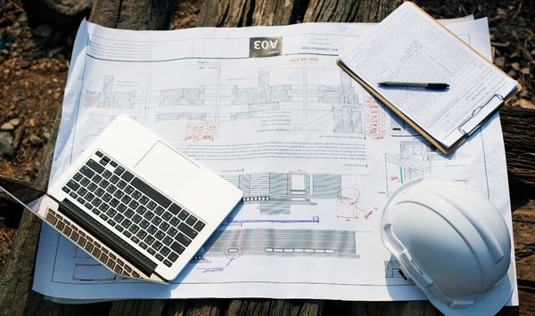 ADU construction plans spread across a table with a laptop, clipboard, pen, and white hard hat.