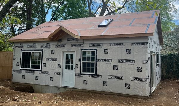 Wood-framed ADU under construction with exterior sheathing, roof rafters, window openings, and a worker on a lift.