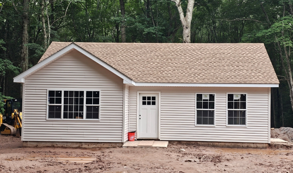 Newly built single-story beige accessory dwelling unit (ADU) with white trim and shingle roof on a wooded residential lot.