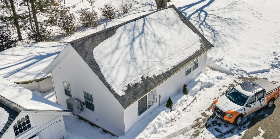 Aerial view of a small light-colored ADU in a snowy yard, with a snow-covered roof, surrounding trees, and a work vehicle parked nearby.