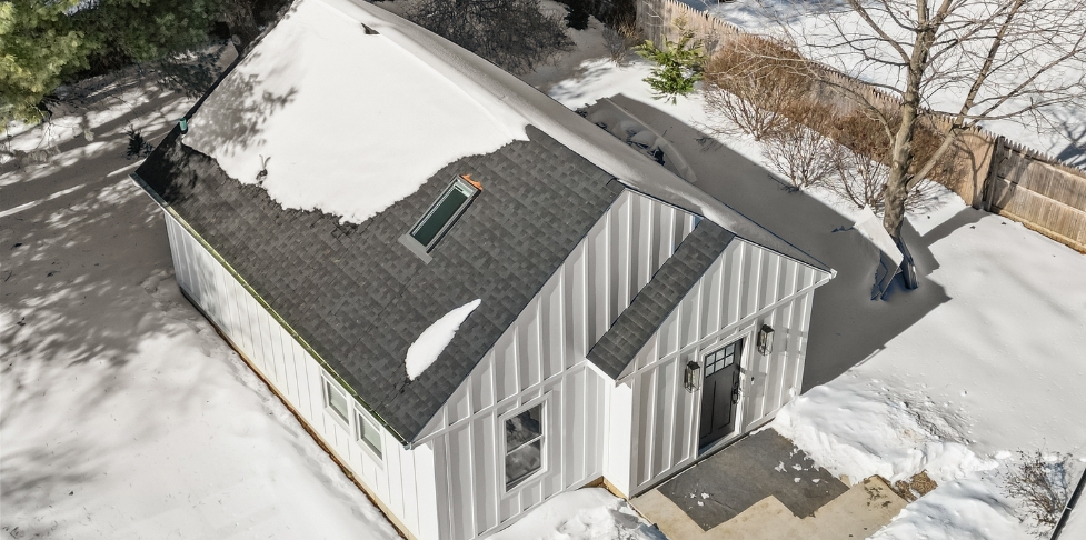 Aerial view of a small white ADU with a dark shingle roof and snow-covered yard, featuring a front gable entry, skylight, and surrounding trees.