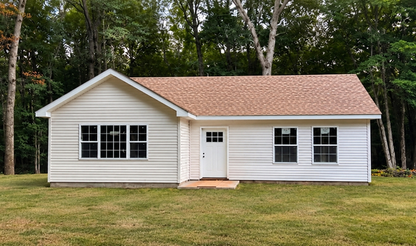 White accessory dwelling unit (ADU) with a red shingle roof, large front windows, and a wooded backyard setting.