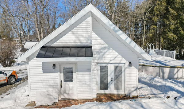 White single-story ADU with a steep gable roof, black metal awning, and snow-covered surroundings in Guilford.