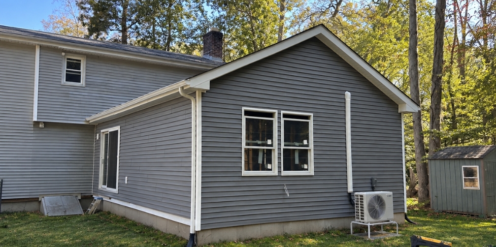 AttachedGray attached ADU addition with white trim, large windows, and an exterior HVAC unit beside a wooded backyard. ADU Example