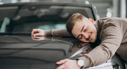 Mann mit legt zufrieden seine rechte Wange auf die Motorhaube eines Autos, von vorne abgebildet