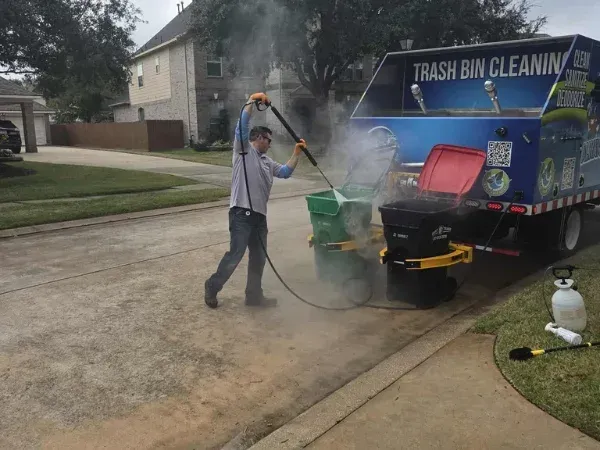 A man in protective eyewear and gloves power washes two trash bins attached to a cleaning truck on a driveway.