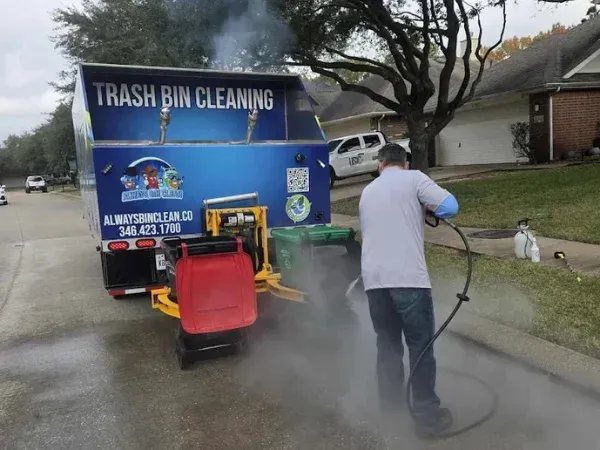 A man in a grey shirt and blue jeans uses a steam cleaner to clean a sidewalk next to a trash bin cleaning truck.