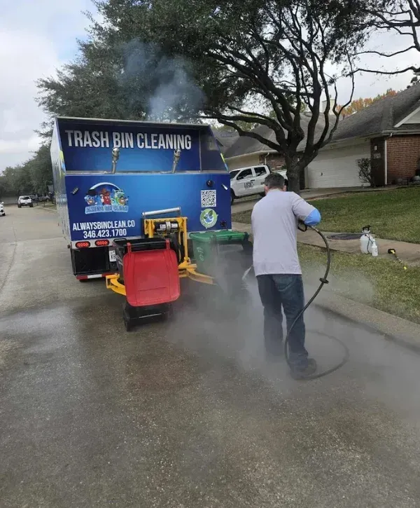 A man in a grey shirt and blue jeans uses a steam cleaner to clean a sidewalk next to a trash bin cleaning truck.