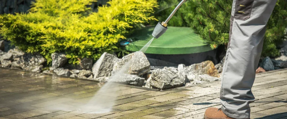 A person in work pants power washing a concrete patio next to green shrubs and rocks on a sunny day.