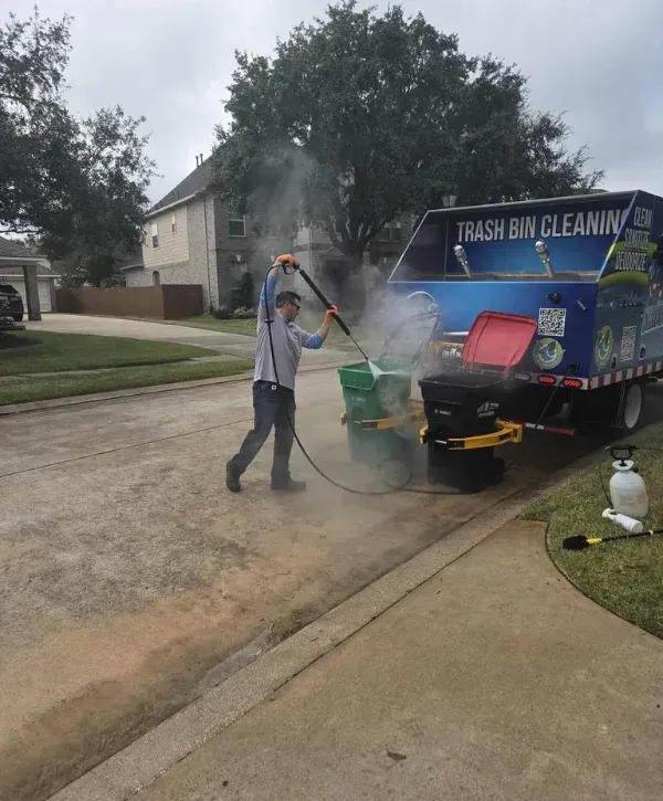 A man in jeans and a gray shirt power washes two trash bins in a residential driveway under an overcast sky.