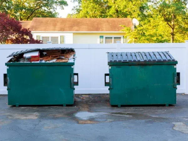 Two large green dumpsters, one overflowing with trash, in front of a white fence and a house.