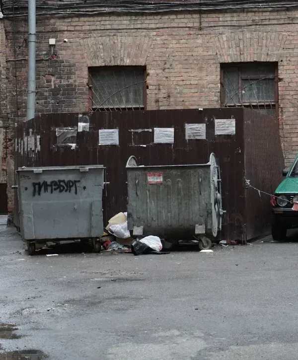 Two large, overflowing dumpsters sit on a grimy concrete ground in an urban back alley, with a brick building and fenced enclosure behind.