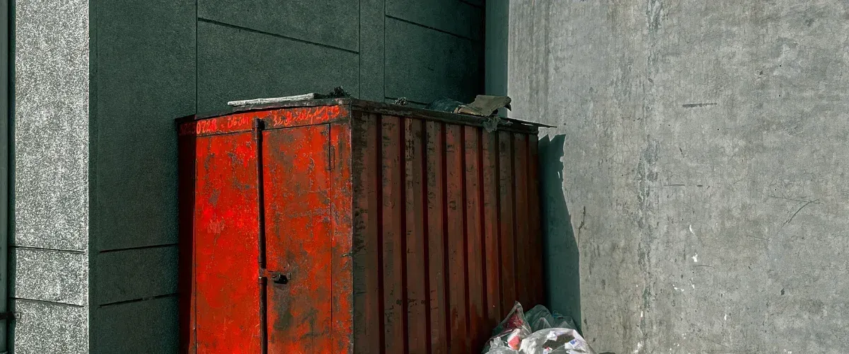 A distressed red metal cabinet with peeling paint next to a textured concrete wall and a dark building facade.