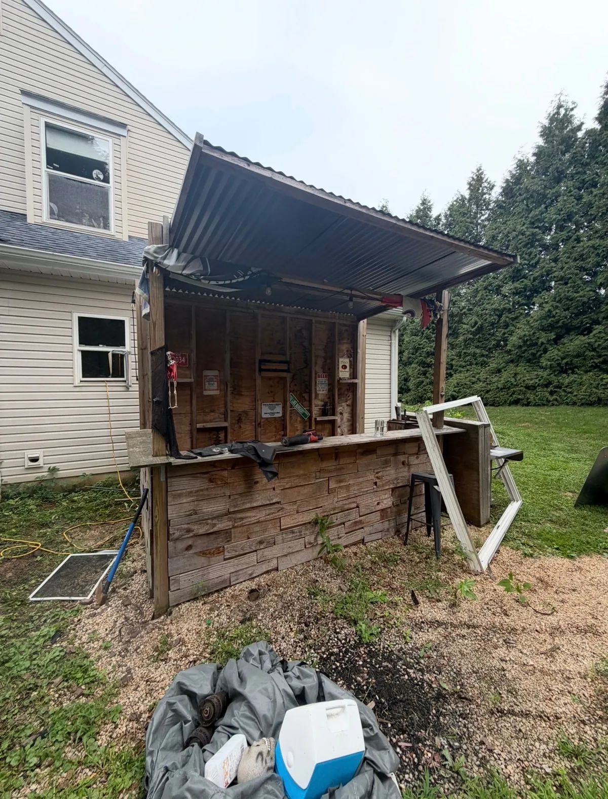 Wooden backyard bar with a corrugated roof, exposed framing, and detached window frames beside a house.